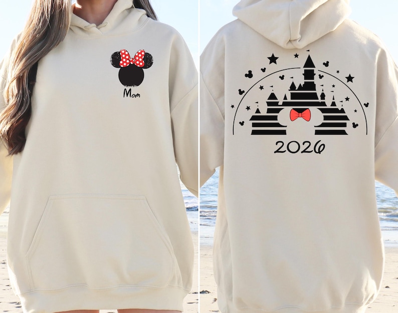 Puede incluir: Sudadera con capucha color crema con bolsillo delantero y capucha. La parte delantera presenta una cabeza de Minnie Mouse negra con un lazo de lunares rojos y blancos y la palabra "Mom" en escritura negra. La parte trasera muestra un dise&ntilde;o de castillo con un lazo rojo y el a&ntilde;o "2026".