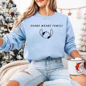 May include: A pale blue sweatshirt featuring the words "OHANA MEANS FAMILY" and a black character outline. The person is wearing light blue jeans and holding a white mug with a Santa Claus design.
