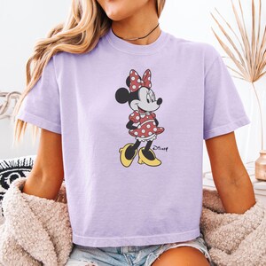 May include: A light purple t-shirt featuring a Minnie Mouse graphic. The cartoon character is wearing a red polka dot dress, bow, and yellow shoes. The Disney logo is printed below the character.