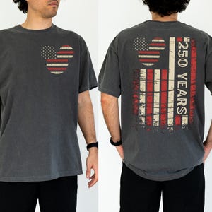 May include: Dark grey t-shirt with a distressed American flag design in the shape of Mickey Mouse ears on the front. The back of the shirt features a larger flag design and the text "250 YEARS".