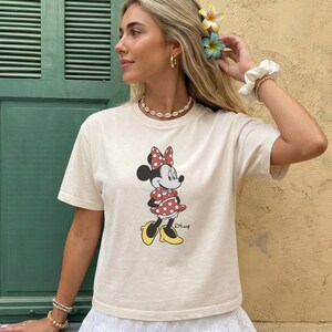 May include: Cream-colored t-shirt featuring a Minnie Mouse graphic. Minnie is depicted in a red polka-dot dress and bow, with yellow shoes. The shirt has short sleeves and a crew neck, suitable for casual wear.