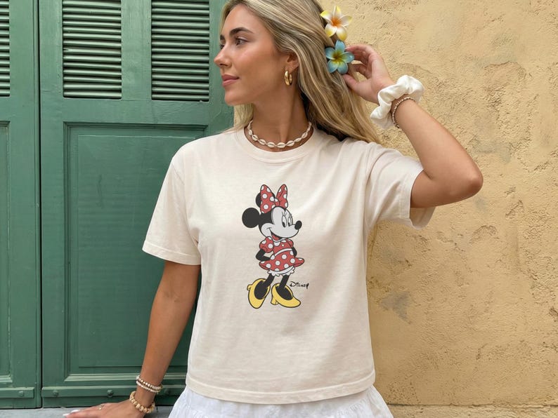 May include: Cream-colored t-shirt featuring a Minnie Mouse graphic. Minnie is depicted in a red polka-dot dress and bow, with yellow shoes. The shirt has short sleeves and a crew neck, suitable for casual wear.