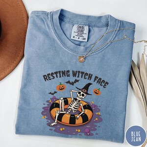 May include: A blue Comfort Colors t-shirt with the phrase "RESTING WITCH FACE" and a skeleton in a witch hat relaxing in a black and orange inner tube. The shirt has a round neckline and short sleeves. A gold necklace is draped across the shirt.