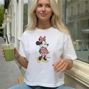 May include: White t-shirt featuring a Minnie Mouse graphic. The cartoon character is wearing a red polka dot dress and bow, with yellow shoes. The shirt is paired with blue jeans. A green drink in a clear cup is held in the hand.