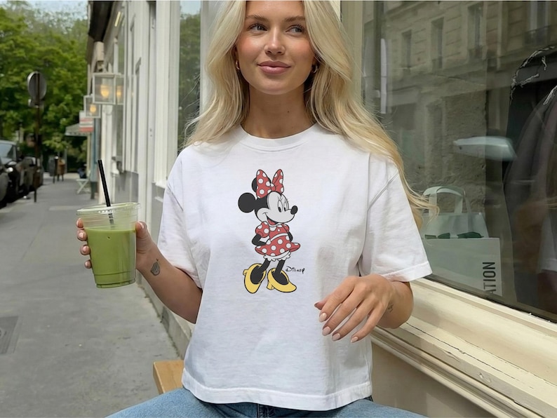 May include: White t-shirt featuring a Minnie Mouse graphic. The cartoon character is wearing a red polka dot dress and bow, with yellow shoes. The shirt is paired with blue jeans. A green drink in a clear cup is held in the hand.