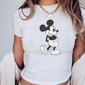 May include: White t-shirt featuring a black and white illustration of Mickey Mouse. The classic cartoon character is depicted in a walking pose, with his signature large ears and smiling face. The shirt is a cropped style.