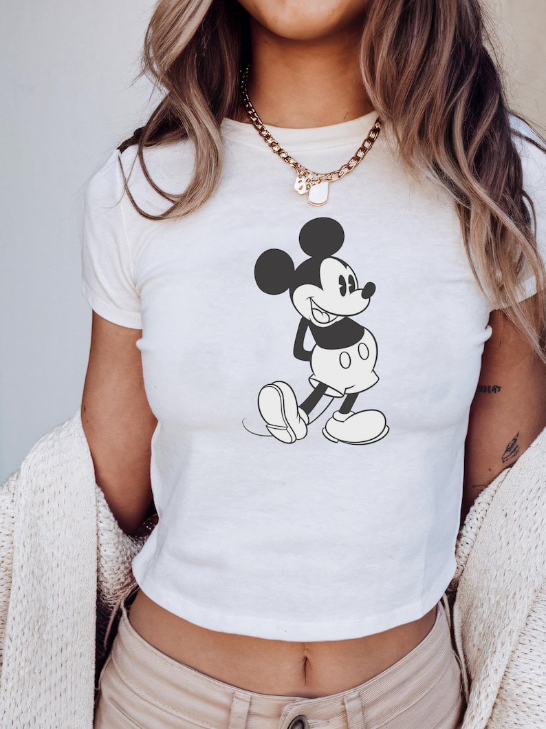 May include: White t-shirt featuring a black and white illustration of Mickey Mouse. The classic cartoon character is depicted in a walking pose, with his signature large ears and smiling face. The shirt is a cropped style.