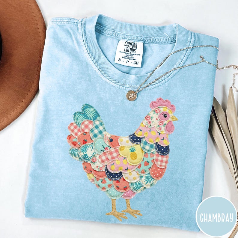 Op de afbeelding: Lichtblauw Comfort Colors T-shirt met een kleurrijk patchwork kippenontwerp. De kip is gemaakt van verschillende stoffen met patronen, waaronder ruitjes, bloemen en fruitprints. Een zilveren ketting met een hanger is ook zichtbaar.