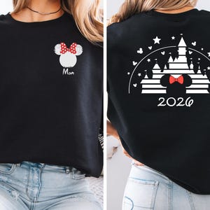 Puede incluir: Sudadera negra con un dise&ntilde;o de Minnie Mouse blanco y un lazo rojo, con la palabra "Mam&aacute;". La parte trasera presenta un dise&ntilde;o de castillo blanco, un lazo de Minnie Mouse y el a&ntilde;o "2026".