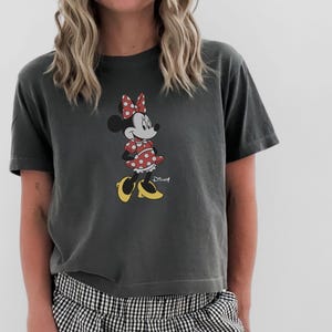 May include: Dark gray t-shirt featuring a Minnie Mouse graphic. Minnie is depicted in her classic outfit: a red polka dot dress, bow, and yellow shoes. The Disney logo is visible.