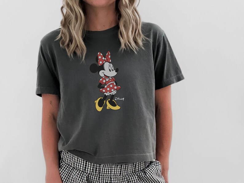 May include: Dark gray t-shirt featuring a Minnie Mouse graphic. Minnie is depicted in her classic outfit: a red polka dot dress, bow, and yellow shoes. The Disney logo is visible.