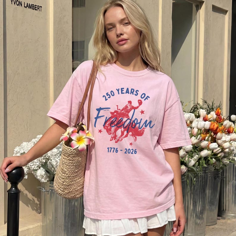 May include: A light pink t-shirt with the text "250 Years of Freedom" and a graphic of a rider on a horse. The shirt also has the dates 1776-2026. The person is wearing a white skirt and carrying a straw bag with flowers.