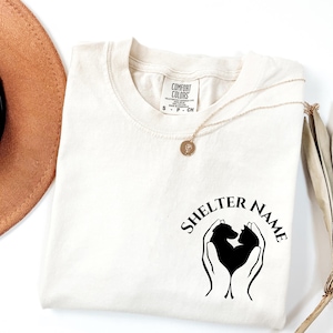 May include: Cream-colored Comfort Colors t-shirt with a black graphic of a dog and cat inside a heart shape, held by two hands. The text "SHELTER NAME" arches above the graphic. A gold necklace and a brown hat are also visible.