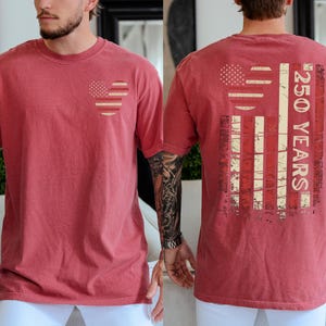 May include: A red t-shirt with a heart-shaped American flag on the front. The back shows a distressed American flag graphic and the text "250 YEARS". The shirt has a vintage appearance. The design is patriotic and commemorates an anniversary.