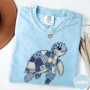 Puede incluir: Camiseta Comfort Colors azul claro con un diseño de tortuga patchwork en varios tonos de azul. La tortuga tiene una cara linda y detalles de estrellas. También se ve un collar plateado con un colgante.