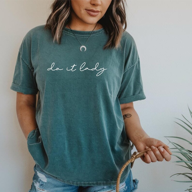 Puede incluir: Camiseta de color verde azulado con la frase "do it lady" en escritura blanca. La camiseta tiene mangas cortas y un corte holgado. La modelo lleva vaqueros azules y un collar plateado.