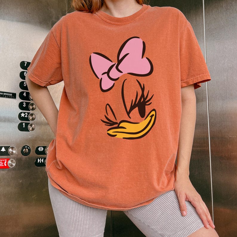 May include: Orange t-shirt featuring a cartoon graphic of a bow, eyelashes, and a duck bill. The graphic is in pink, black, and yellow. The shirt is a short-sleeved, crew neck style.