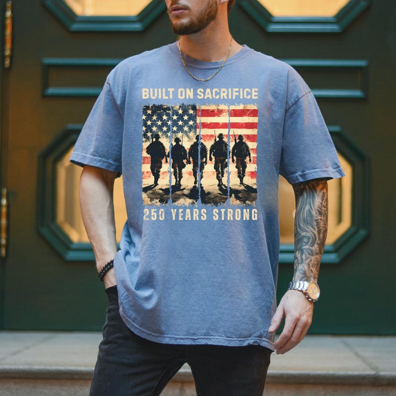 May include: Blue t-shirt with the text "BUILT ON SACRIFICE" and "250 YEARS STRONG." The shirt features an American flag design with silhouettes of soldiers. The shirt is a comfortable, casual style.