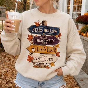 May include: A cream-colored crewneck sweatshirt featuring a graphic of a wooden signpost with four directional arrows. The signs read Stars Hollow, Dragonfly Inn, Luke's Diner with a coffee cup icon, and Kim's Antiques, all surrounded by autumn leaves.