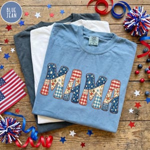 May include: Three t-shirts in blue, white, and gray are stacked on a wooden surface. The top shirt is light blue with the word "MAMA" in a patchwork design of red, white, and blue stars and patterns. Patriotic decorations surround the shirts.