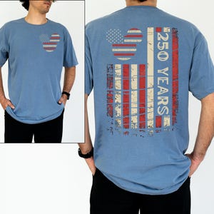 May include: A blue short-sleeved t-shirt with a distressed American flag design on the front and back. The front features a Mickey Mouse-inspired flag, while the back displays a vertical flag design with the text "250 YEARS".