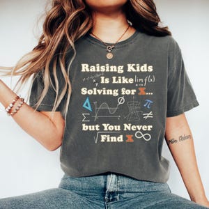 May include: Dark gray t-shirt with the text "Raising Kids Is Like Solving for X... but You Never Find X" in white and orange. The shirt features mathematical symbols and equations, including the limit of f(x), pi, and infinity.