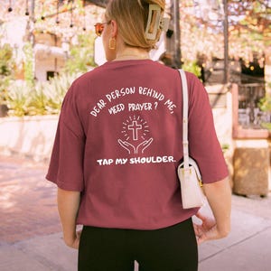 May include: A maroon t-shirt with white text that reads "Dear person behind me, need prayer? Tap my shoulder." The text is accompanied by a graphic of a cross with hands reaching up to it.