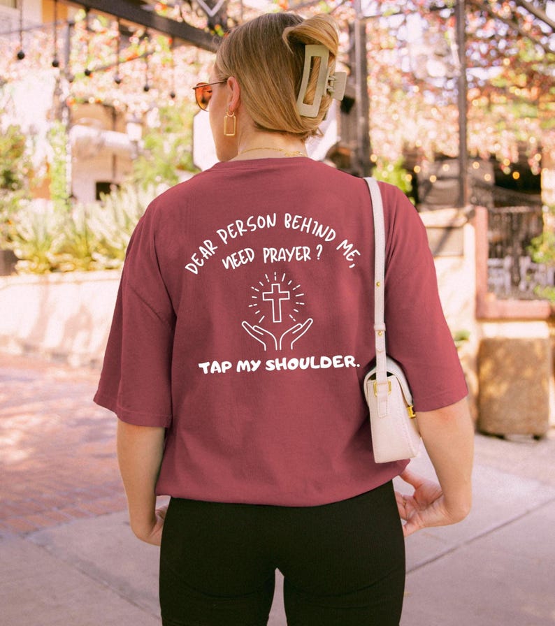 May include: A maroon t-shirt with white text that reads "Dear person behind me, need prayer? Tap my shoulder." The text is accompanied by a graphic of a cross with hands reaching up to it.