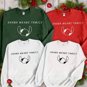 May include: Four sweatshirts in green, red, and white, each with the text "OHANA MEANS FAMILY" and a cartoon character outline. Festive holiday decorations surround the shirts, including red baubles and greenery.