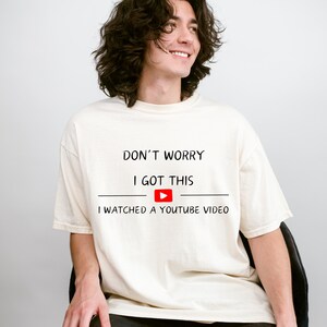 May include: Cream-colored t-shirt with the text "DON'T WORRY I GOT THIS" above a YouTube play button and the phrase "I WATCHED A YOUTUBE VIDEO". The shirt is a casual, unisex style.