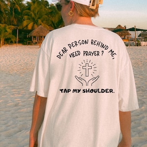 May include: White t-shirt with black text that reads "Dear person behind me, need prayer? Tap my shoulder." and a black cross with hands reaching up to it.