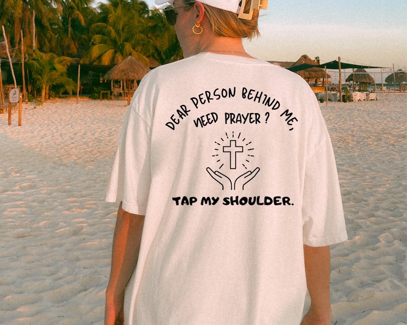 May include: White t-shirt with black text that reads "Dear person behind me, need prayer? Tap my shoulder." and a black cross with hands reaching up to it.