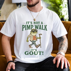 May include: White t-shirt with the text "IT'S NOT A PIMP WALK IT'S GOUT!" and a cartoon frog wearing sunglasses, a hat, and holding a cane. The frog is dressed in a plaid shirt, orange shirt, blue jeans, and brown shoes.