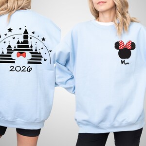 Puede incluir: Sudadera azul claro con un dise&ntilde;o de castillo negro, un lazo rojo y el a&ntilde;o 2026 en la espalda. La parte delantera muestra una cabeza de Minnie Mouse negra con un lazo de lunares rojos y la palabra "Mom" en negro.