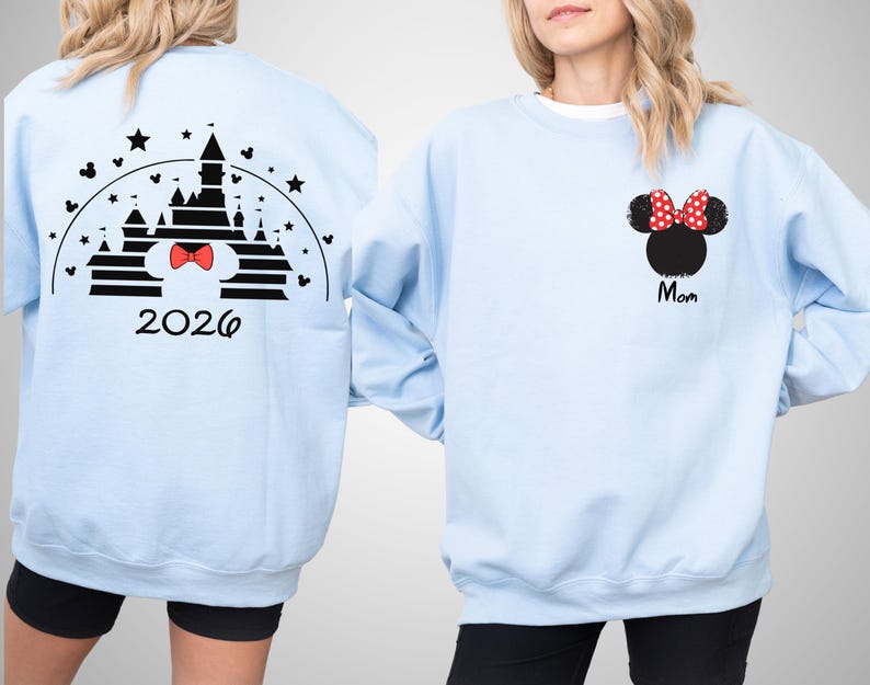 Puede incluir: Sudadera azul claro con un dise&ntilde;o de castillo negro, un lazo rojo y el a&ntilde;o 2026 en la espalda. La parte delantera muestra una cabeza de Minnie Mouse negra con un lazo de lunares rojos y la palabra "Mom" en negro.