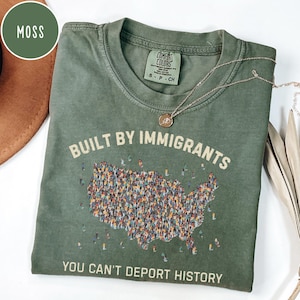 May include: A moss green t-shirt with the text "BUILT BY IMMIGRANTS" above a graphic of the United States filled with tiny figures. The phrase "YOU CAN'T DEPORT HISTORY" is printed below. A gold necklace rests on the shirt.