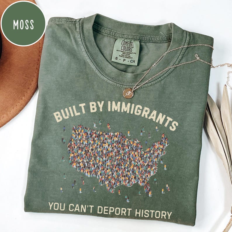 May include: A moss green t-shirt with the text "BUILT BY IMMIGRANTS" above a graphic of the United States filled with tiny figures. The phrase "YOU CAN'T DEPORT HISTORY" is printed below. A gold necklace rests on the shirt.