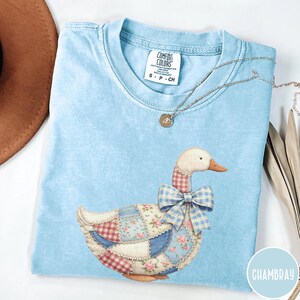 Puede incluir: Camiseta azul claro Comfort Colors con un diseño de ganso patchwork y un lazo de cuadros. El ganso tiene una cabeza blanca, un pico naranja y un cuerpo hecho de varias telas estampadas. También se ve un collar plateado con un colgante.