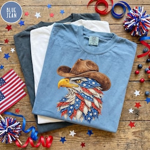 May include: Three t-shirts in gray, white, and light blue are stacked on a wooden surface. The top shirt is light blue and features a graphic of an eagle wearing a cowboy hat with an American flag design. Patriotic decorations surround the shirts.