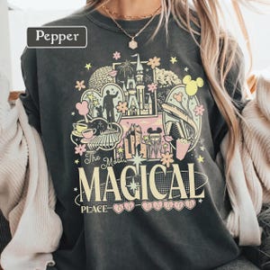 May include: A charcoal gray crewneck sweatshirt featuring a graphic print with the text "The Most MAGICAL PLACE ON EARTH" and "Pepper". The design includes illustrations of a castle, a monorail, a teacup, and stars in shades of pink, yellow, and white.