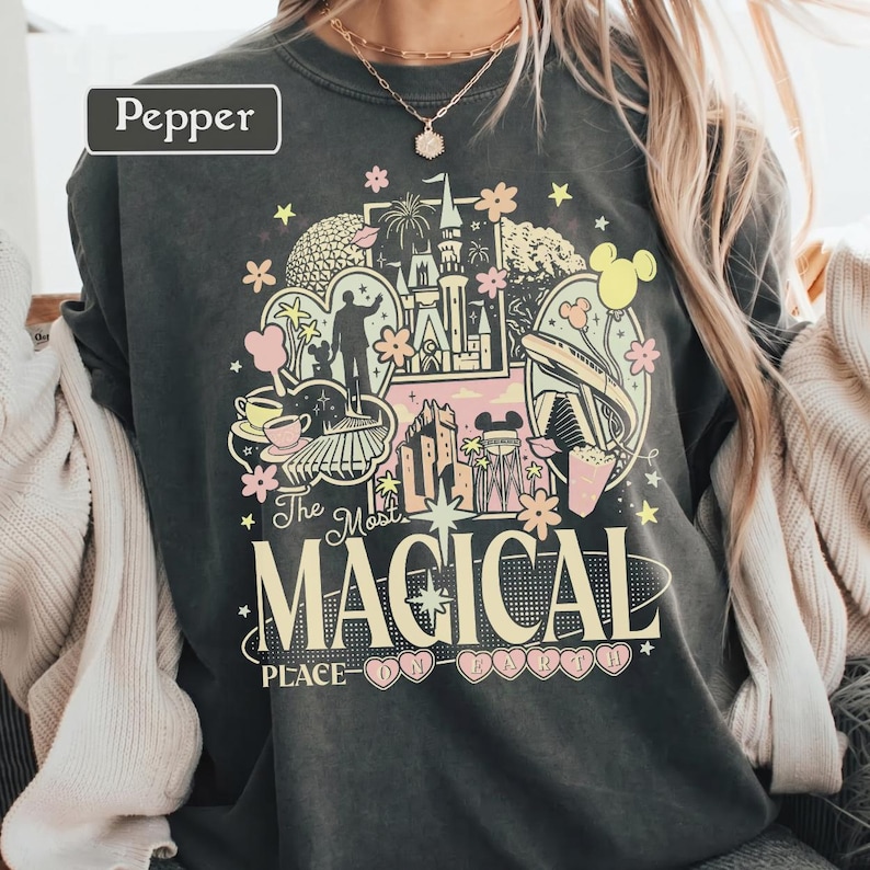 May include: A charcoal gray crewneck sweatshirt featuring a graphic print with the text "The Most MAGICAL PLACE ON EARTH" and "Pepper". The design includes illustrations of a castle, a monorail, a teacup, and stars in shades of pink, yellow, and white.