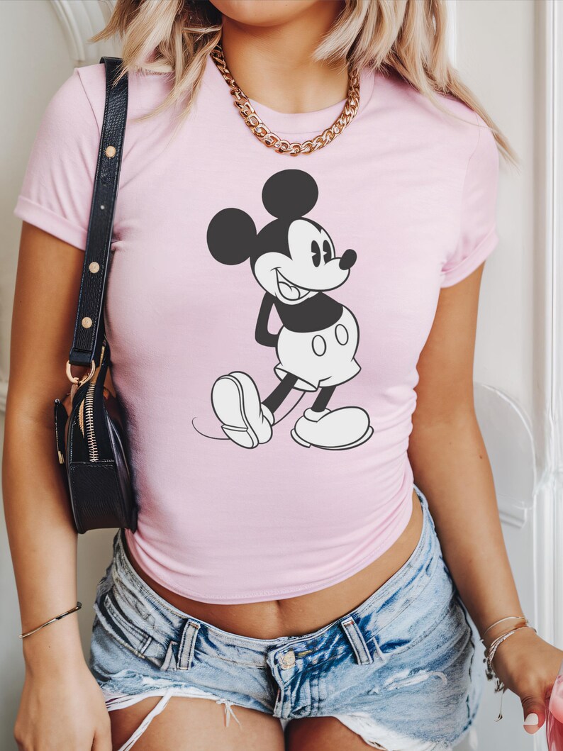 May include: A light pink t-shirt featuring a black and white graphic of Mickey Mouse. The cartoon character is in a classic pose. The shirt has short sleeves.