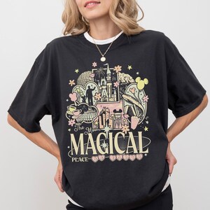 May include: A black short-sleeve t-shirt featuring a graphic print with various theme park icons, including a castle, a monorail, and teacups. The text reads "The Most MAGICAL PLACE ON EARTH" in a mix of serif and sans-serif fonts with floral accents.