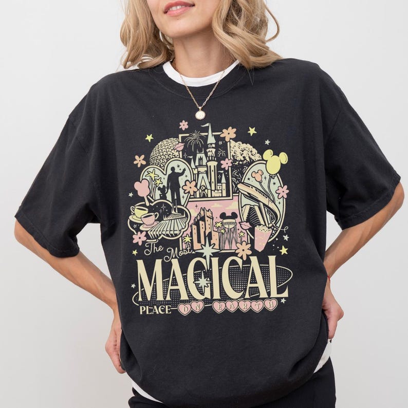 May include: A black short-sleeve t-shirt featuring a graphic print with various theme park icons, including a castle, a monorail, and teacups. The text reads "The Most MAGICAL PLACE ON EARTH" in a mix of serif and sans-serif fonts with floral accents.