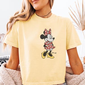May include: Yellow t-shirt featuring a graphic of Minnie Mouse. Minnie is depicted in her classic outfit: a red polka-dot dress, matching bow, and yellow shoes. The Disney logo is visible below the character.
