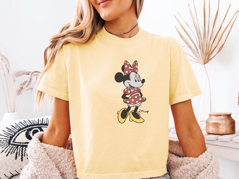 May include: Yellow t-shirt featuring a graphic of Minnie Mouse. Minnie is depicted in her classic outfit: a red polka-dot dress, matching bow, and yellow shoes. The Disney logo is visible below the character.