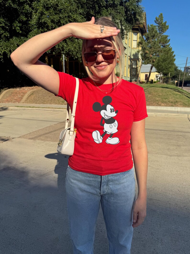 May include: A red t-shirt featuring a classic Mickey Mouse graphic. The shirt is paired with light blue jeans and a small, cream-colored purse. The person is wearing sunglasses and standing outside on a sunny day.