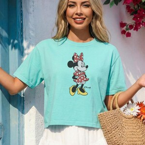 May include: A light turquoise t-shirt featuring a Minnie Mouse graphic. The cartoon character is wearing a red polka dot dress, bow, and yellow shoes. A woven straw tote bag with flowers is also visible.
