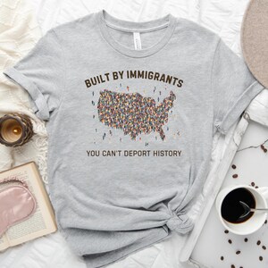 May include: A light gray t-shirt with the text "BUILT BY IMMIGRANTS" above a map of the United States filled with tiny figures. Below the map, the text reads "YOU CAN'T DEPORT HISTORY."