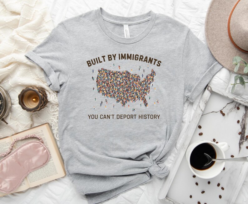 May include: A light gray t-shirt with the text "BUILT BY IMMIGRANTS" above a map of the United States filled with tiny figures. Below the map, the text reads "YOU CAN'T DEPORT HISTORY."